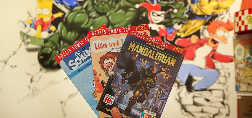 Eine Auswahl der Gratis Comics am Aktionstag Vor einer Wand mit Superheldenzeichnungen werden drei Comichefte präsentiert.