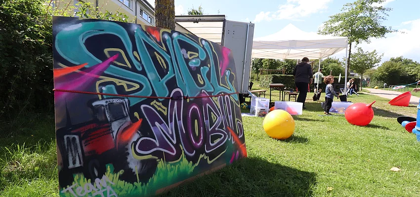 Das Spielmobil-Team sorgt für schöne Nachmittage Kinder spielen auf einer Wiese, dafür ein Schild mit der Aufschrift "Spielmobil"