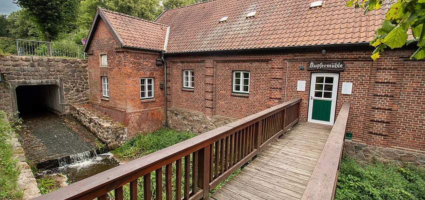 Kupfermühle Glinde (2024) Ein Backsteingebäude mit Satteldach (Kupfermühle) mit Holzbrücke über die Glinder Au.