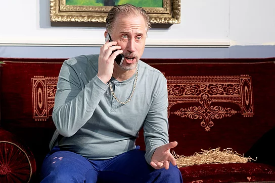 Schotty aus "Der Tatortreiniger" Ein Mann mit Arbeitshose sitz auf einer Couch und telefoniert überrascht.